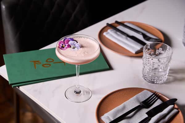 A vibrant cocktail garnished with edible flowers, served in a stylish glass on a table set with plates and utensils, showcasing the elegant ambiance of the Indian restaurant.