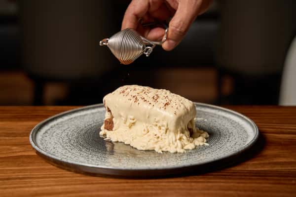 A hand drizzling sauce over a decadent dessert of layered cake topped with creamy frosting, elegantly presented on a textured plate, highlighting the artistry of dessert preparation.
