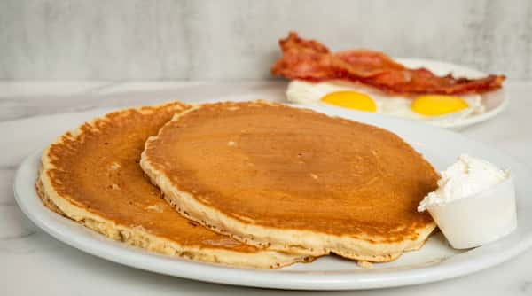 Stack of fluffy golden pancakes served with crispy bacon and eggs on a white breakfast plate.