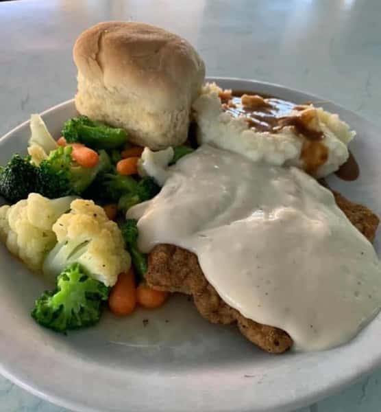 Fried chicken topped with gravy and a side of mashed potatoes, vegetables, and a biscuit