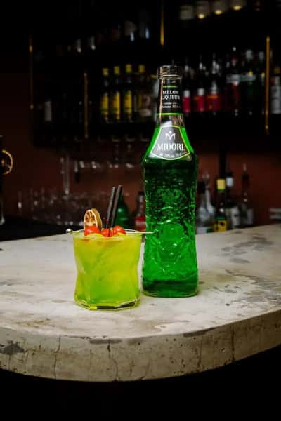 Bright green cocktail with citrus garnish served beside a tall glass bottle on a marble bar counter