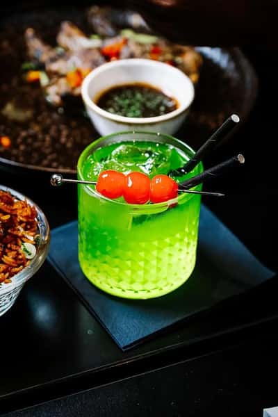 Bright neon green cocktail garnished with skewered cherries, served in a textured glass on a bar table with small plates and savory dishes in the background