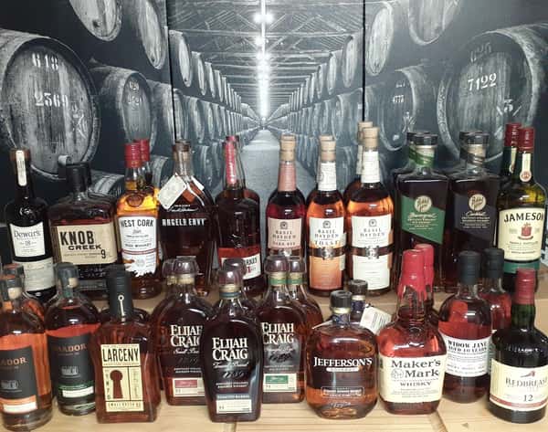 Extensive collection of premium whiskey and bourbon bottles, including Elijah Craig, Basil Hayden, Knob Creek, and Jameson, displayed in front of a black-and-white barrel warehouse mural, ideal for a whiskey bar or tasting room setting.