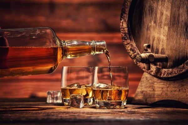 Close-up of golden whiskey being poured from a bottle into two glasses with ice cubes, set against a rustic wooden barrel and warm-toned background, evoking a cozy, craft distillery atmosphere.