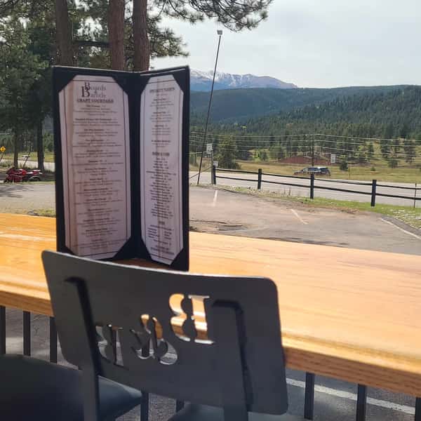 Craft cocktail menu from Boards & Barrels displayed on a wooden outdoor table with scenic views of pine-covered hills and snow-capped mountains, offering a relaxed, mountain-side dining experience.