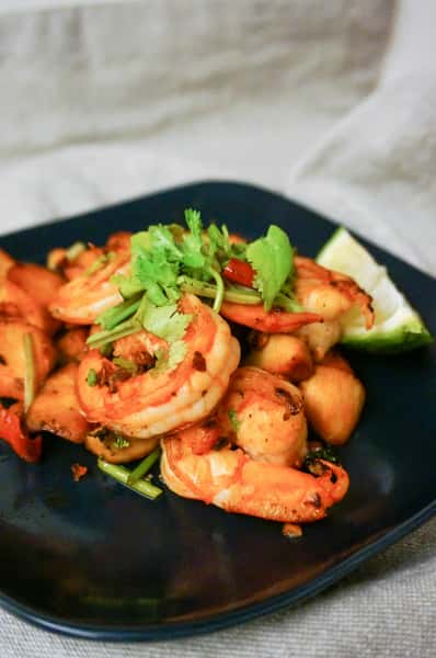 Sautéed shrimp with golden potatoes, red peppers, and fresh cilantro, served with a lime wedge on a dark plate.