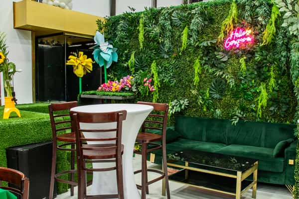 Lush green photo-op area featuring a faux foliage wall, a neon "Hello Gorgeous" sign, velvet green couch, and playful decor including oversized pinwheels and floral arrangements.