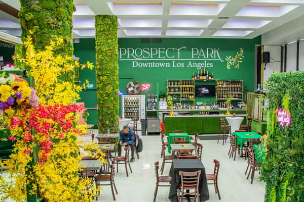 This vibrant and lush venue, Prospect Park Downtown Los Angeles, features a nature-inspired aesthetic with green faux grass, foliage-covered columns, and colorful floral arrangements.