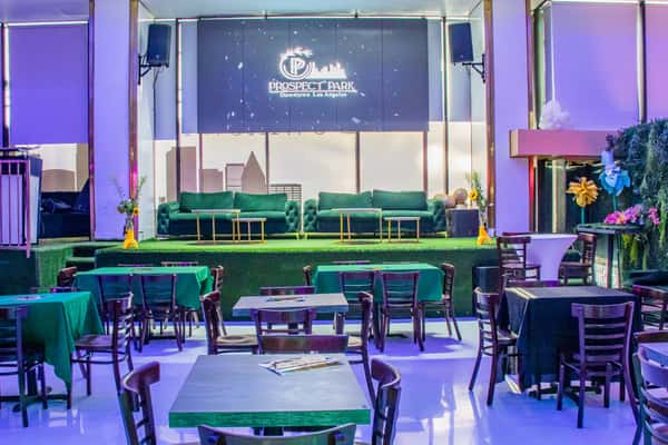 Colorful lounge with green sofas on stage, wooden tables, and Prospect Park Downtown Los Angeles sign lit in the background.