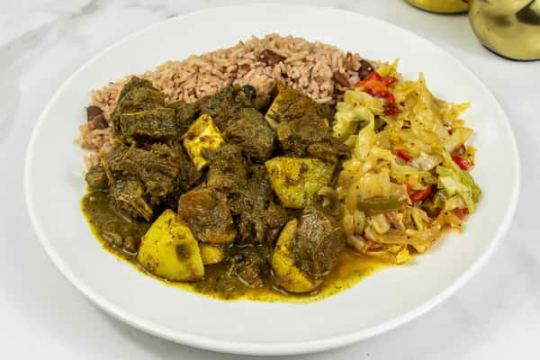 Jamaican curry goat with potatoes, served with rice and peas and sautéed cabbage on a white plate.