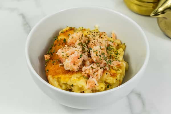 Baked macaroni and cheese topped with seasoned shrimp and herbs served in a white bowl on a marble surface.