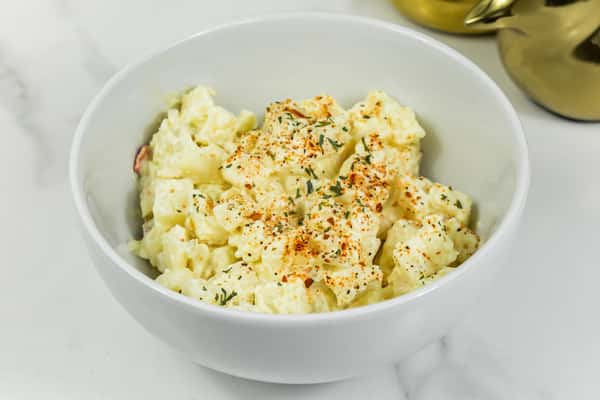Creamy potato salad served in a white bowl, topped with paprika and fresh herbs.