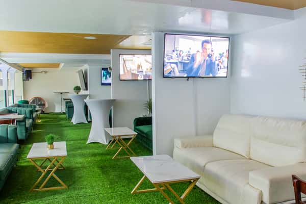 Modern lounge area with green turf flooring, white and green sofas, marble-top tables, and multiple wall-mounted flat-screen TVs.