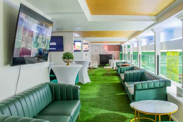 Upscale indoor lounge with green leather sofas, grass-like carpet, cocktail tables, and wall-mounted TVs in a bright setting.
