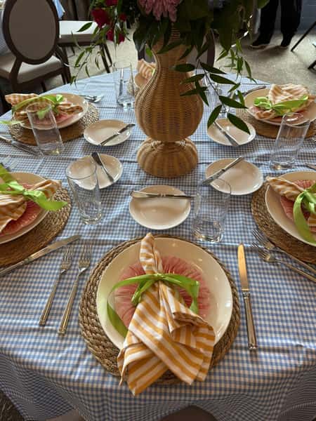 Charming private dining table set with blue gingham tablecloth, woven placemats, striped napkins tied with green ribbon, and a floral centerpiece, styled for an intimate restaurant event or catered gathering