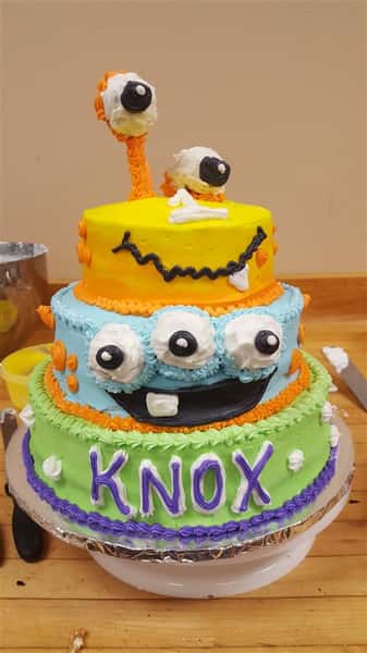 Three layer green, blue and yellow cake decorated with a different monster at each layer with"Know" written on the bottom layer