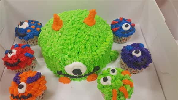 frosted cake resembling a green and orange monster with one eye surrounded by cupcakes that are frosted to resemble various monsters