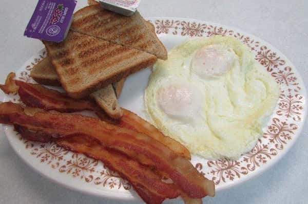 Plate with sunnyside up eggs and three pieces of bacon with a side of toast and grape jelly
