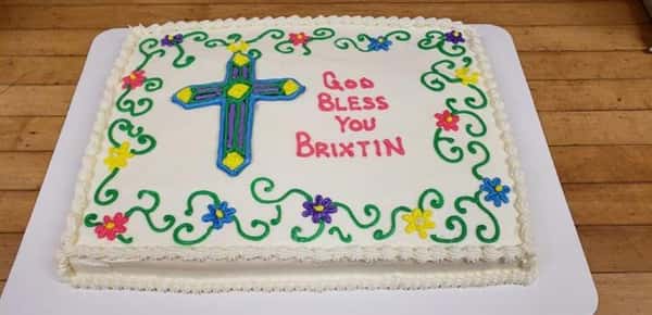 White rectangle cake that says God Bless You Brixtin decorated with flowers and a cross in frosting