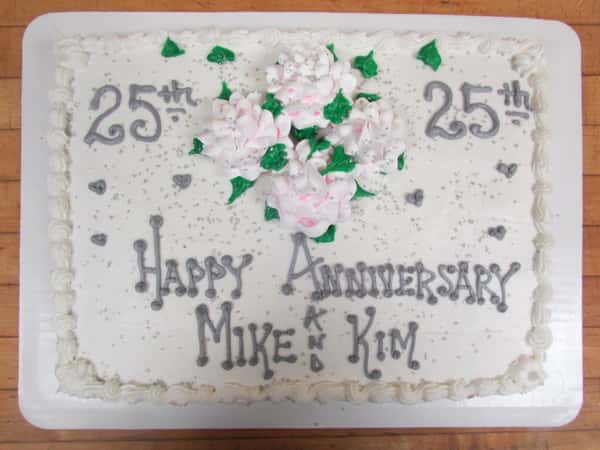 White cake with Happy 25th Anniversary Mike and Kim on it in silver decorated with four light pink flowers in the center