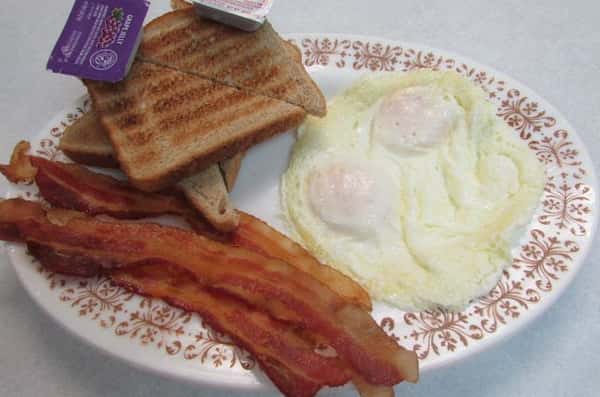 Plate of three sunny side up eggs, three slices of bacon, and toasted bread cut in half with a side of grape jelly