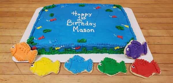A blue rectangle cake that is decorated like an ocean that sayas Happy 1st Birthday Mason with three fish shaped cookies next to it