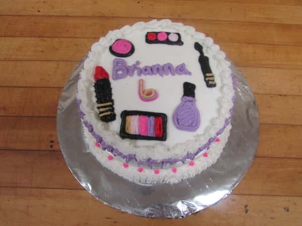 White frosted cake that reads " Brianna 6" decorated with lipstick, eye shadow, nail polish, and a mascara wand made of frosting
