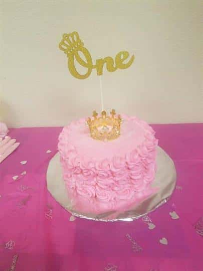 Pink frosted cake with a gold crown on top a a gold decoration that says "One" on it