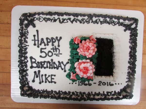 White square cake that reads "Happy 50th Birthday Mike 1966-2016" decorated with frosted flowers and a white squiggly border