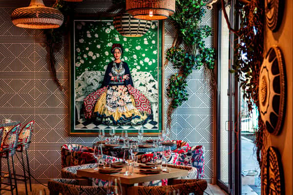 Stylish restaurant dining area with bold geometric wallpaper, colorful upholstered seating, hanging woven lights, and large Frida Kahlo portrait