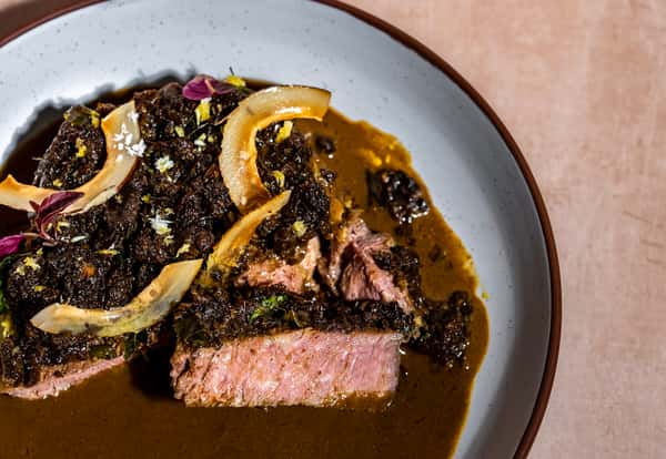 Medium-rare sliced steak in rich brown sauce with caramelized onions, herbs, and citrus garnish in a modern fine dining bowl