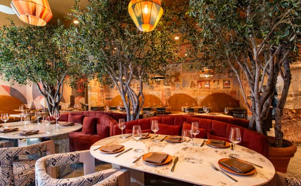 Elegant upscale restaurant dining room with marble tables, velvet banquette seating, indoor olive trees, and warm ambient lighting
