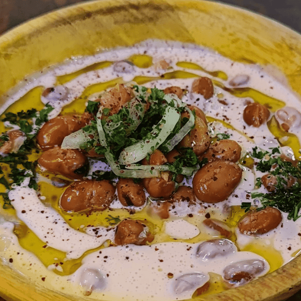Creamy fava beans drizzled with olive oil, topped with parsley, onions, and spices, served in a rustic wooden bowl.