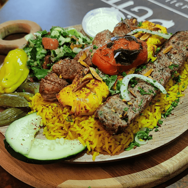 Mixed grill platter with beef kebabs, chicken skewers, grilled tomato, yellow rice, pickles, and a side of fresh salad and garlic sauce.