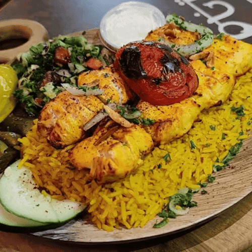 Grilled chicken kebabs served on yellow rice with charred tomato, fresh salad, pickles, and a side of creamy dipping sauce.