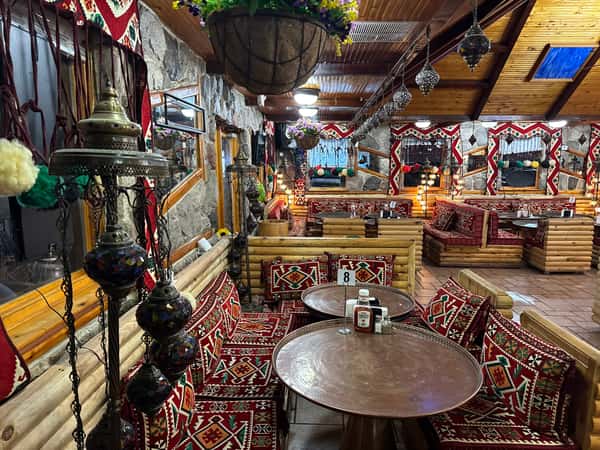 Cozy Mediterranean restaurant interior with wooden log seating, vibrant red patterned cushions, and colorful hanging mosaic lamps.