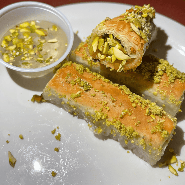 Flaky pistachio-filled baklava rolls drizzled with syrup, served with a side of crushed pistachio syrup for dipping.