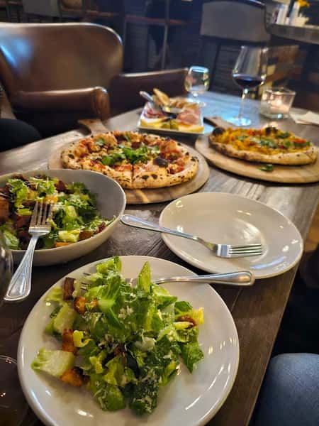 Plates of fresh Caesar salad served alongside wood-fired pizzas, with wine glasses and charcuterie on a rustic wooden table.