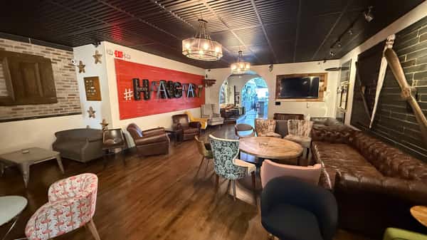Stylish lounge with mixed seating, round wooden tables, a leather sofa, and a bold red wall featuring "#HANGAR7" signage.