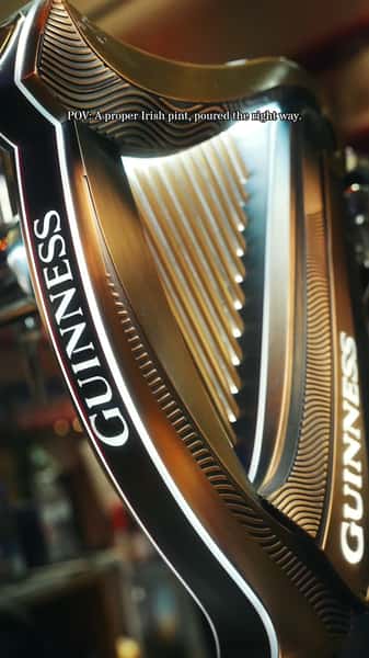Close-up of a Guinness harp beer tap handle with warm bar lighting and the caption "POV: A proper Irish pint, poured the right way."
