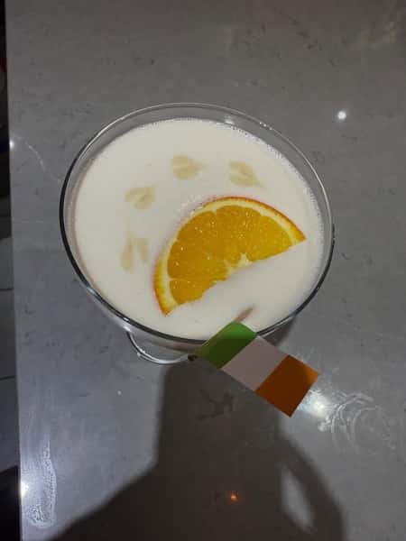 Creamy cocktail topped with foam, garnished with an orange slice and a small Irish flag on the rim, served in a coupe glass on a marble bar counter