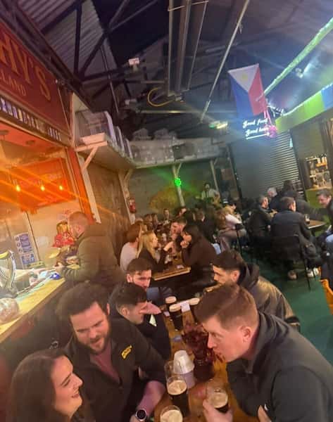 Crowded bar or food hall with groups of people sitting at long tables drinking beer and socializing under hanging lights and flags.