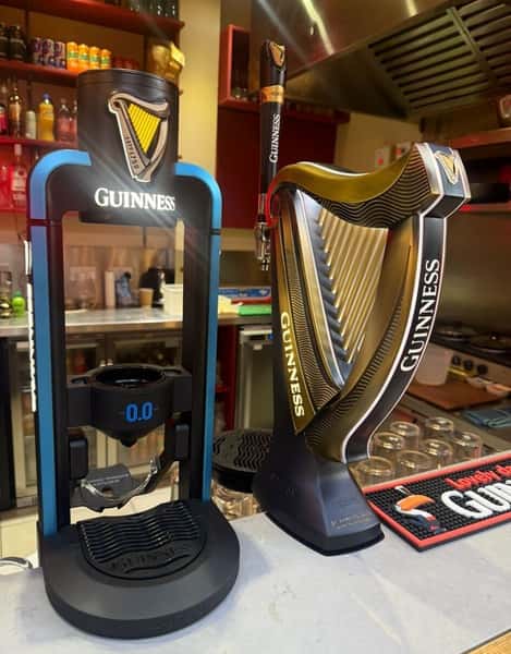 Two Guinness beer taps on a bar counter—one with a harp-shaped design and the other with a digital display—set in front of shelves with bottles and bar equipment.