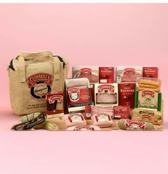 A promotional display of various packaged Clonakilty food products, including bacon rashers, sausages, black pudding, and other meats, arranged with a branded tote bag against a pink background.
