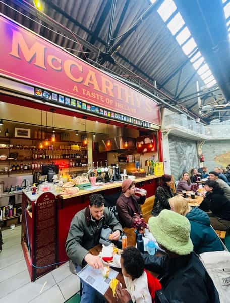 A busy food stall called "McCarthy's – A Taste of Ireland" inside an indoor market, with a bar counter serving drinks and food while groups of people sit at long tables eating, drinking, and socializing.