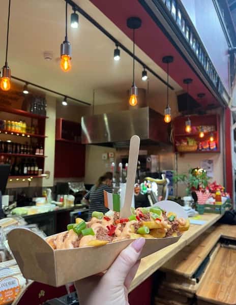 A hand holding a tray of loaded fries topped with sauce and chopped green onions, in front of a food stall kitchen with hanging lights and a wooden bar counter.
