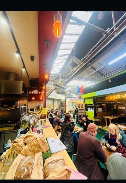 Indoor food market or food hall with people sitting at tables, a bar counter with beer taps and bread on display, and small restaurant stalls in the background.