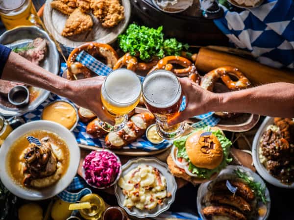 Two people hold glasses of beer over a table filled with different german foods.
