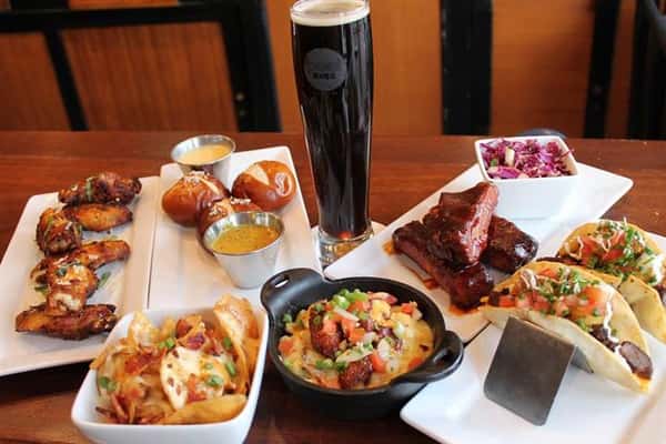 a plate of pretzel bites with 2 dipping sauces, a bowl of nachos, and plate of ribs with a side salad, and a bow of chopped vegetables and meat.