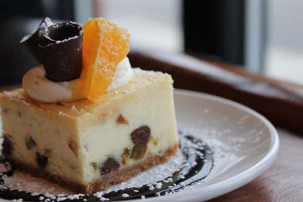 a piece of cheesecake with cream, chocolate, and a peeled orange slice on top